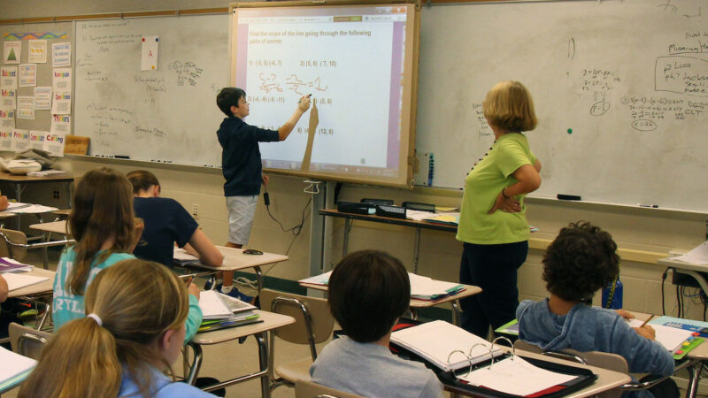 Students and a teacher watch as a classmate answers a math question on the white board.