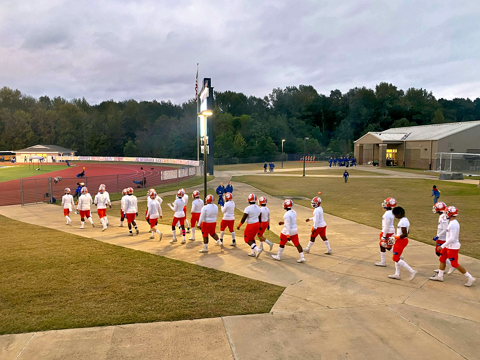 The Madison Central Jaguars football team, wearing white jerseys and orange pants, marches down to the field in Jaguar Stadium ahead of a Friday night football game against Murrah High School in Mississippi. Madison Central High School does not require a COVID vaccine for student athletes to participate in sports, while Murrah High School does.