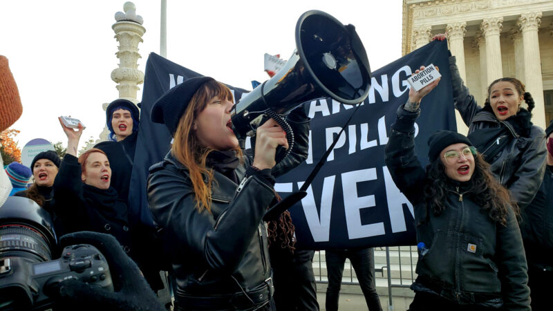 A woman who is part of a group of abortion-rights advocates rallying outside of the U.S. Supreme Court yells into a microphone while other demonstrators hold up small, white signs that say "abortion pills" on them.