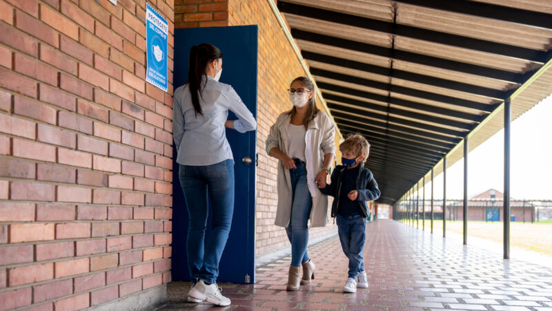 Teacher welcoming a student with her mother back to school and wearing facemasks