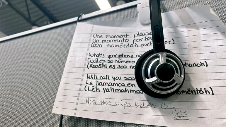 A note at a desk in the ViaLink office in New Orleans Louisiana lists translations for phrases workers might use during a mental health hotline card, including: "One moment please," "What's your phone number?" and "We'll call you soon."