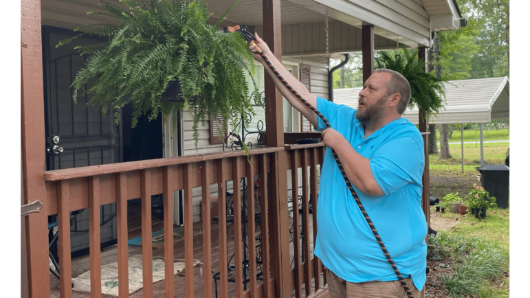 Man in a blue shirt waters a fern hanging in front of his house.