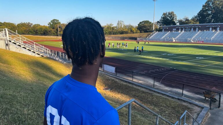 A boy in a jersey stands on the stairs looking out onto a football field.