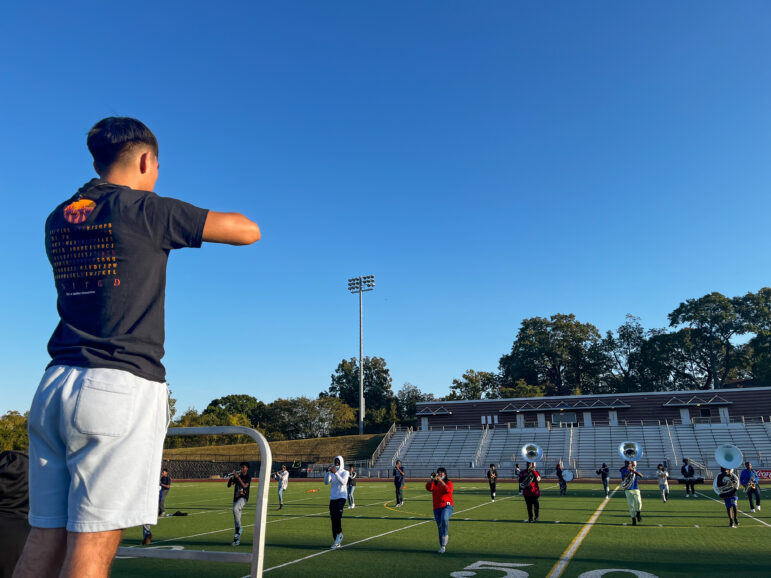 A young man stands on a lift on a football field directing the band practice