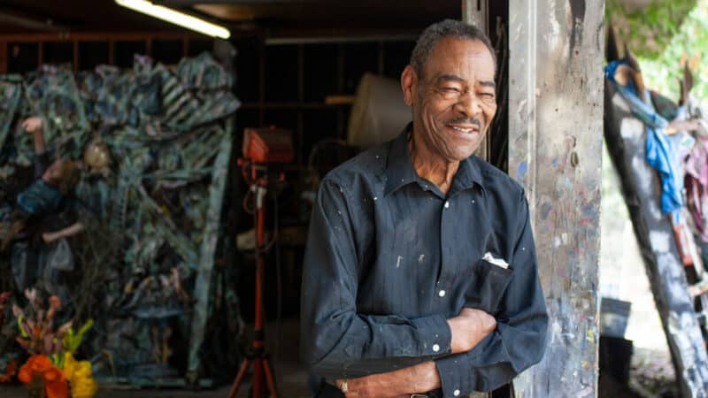 The artist Thornton Dial poses, smiling, outside of his studio.