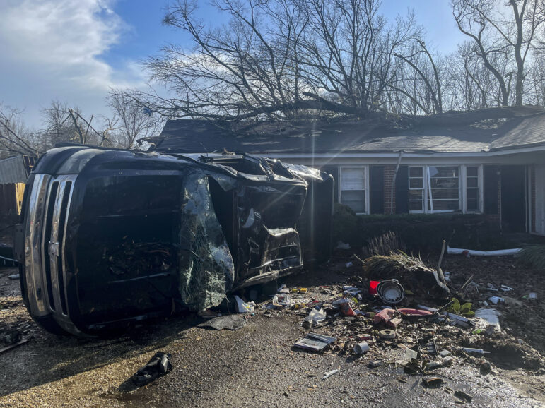 A car rest on its side with its windshield busted out in front of a home with a large tree limb on its roof. Debris is scattered throughout the yard.