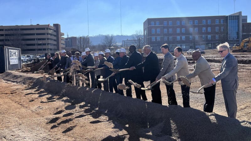 A line of people in hard hats and suits shovel dirt during a groundbreaking ceremony.