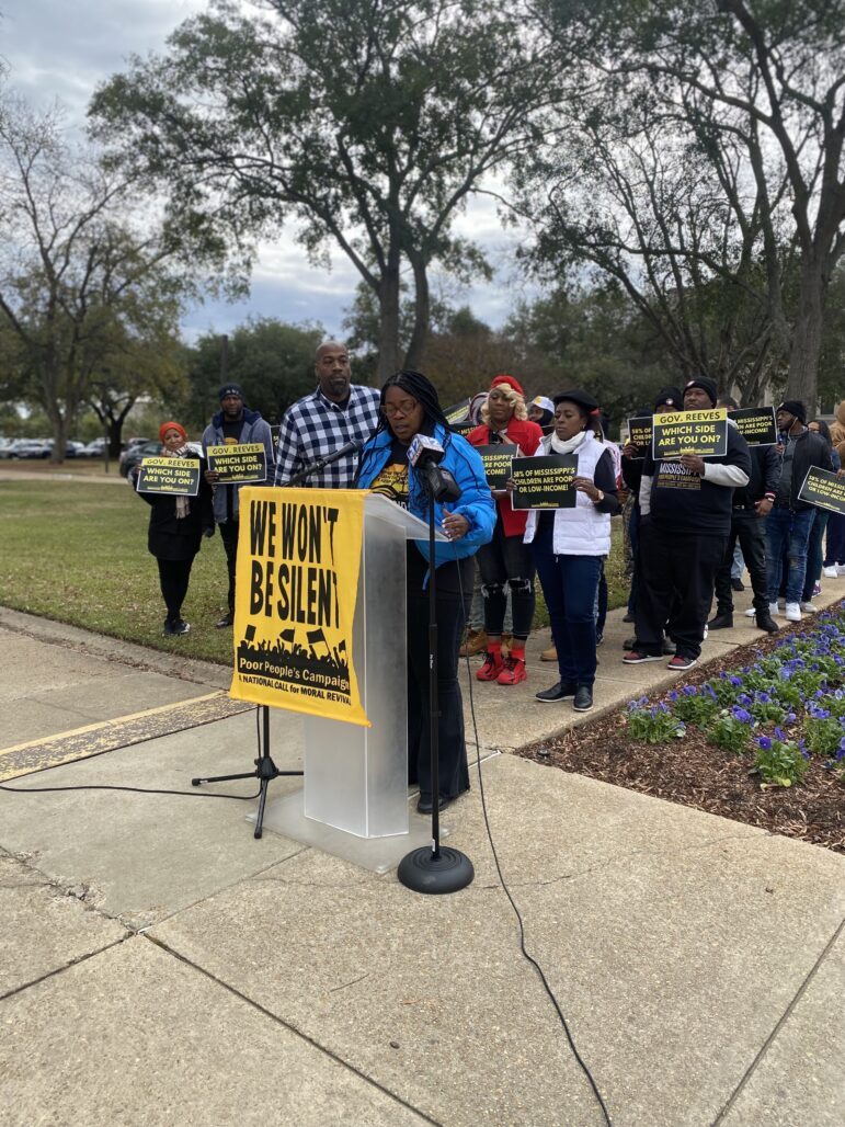 An African American woman speaks behind a podium with the sign "WE WON'T BE SILENT."