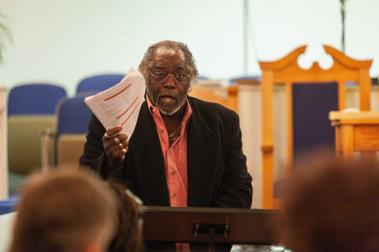 An African American man speaks while holding a stack of documents.