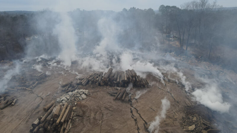 Smoke rises from an area made up of dirt and piles of logs.