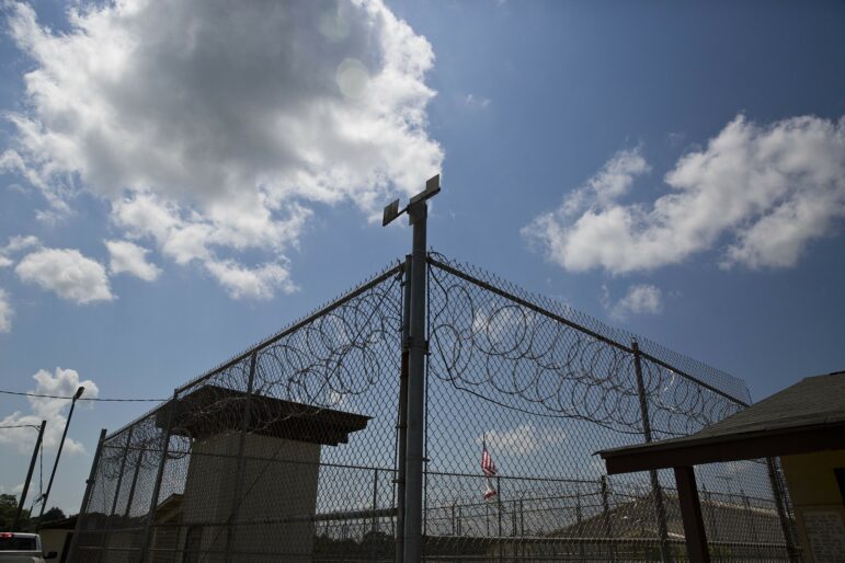 A chainlink fence with barbed wire and a guard tower in the background stands against a blue sky with some clouds.