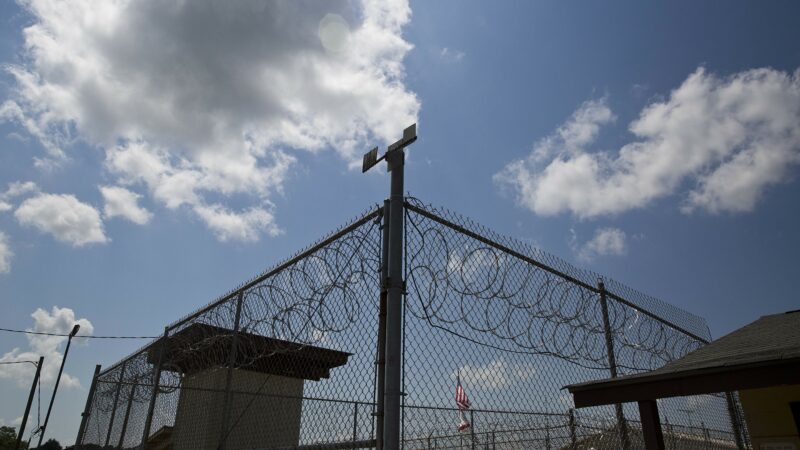 A chainlink fence with barbed wire and a guard tower in the background stands against a blue sky with some clouds.