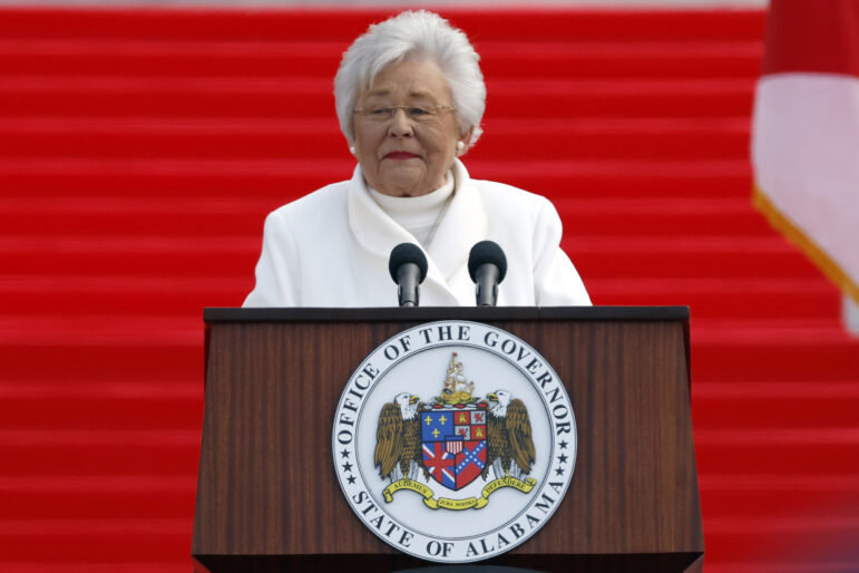 Gov Kay Ivey speaks at a podium in front of a red background.