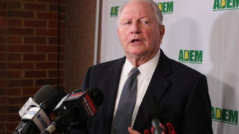 An older white man speaks into microphones in front of a background with the ADEM logo on it.