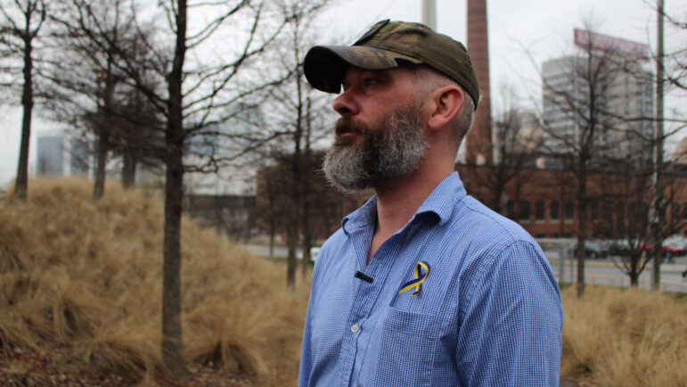 A man in a camo hat and a blue and yellow ribbon pinned to his shirt looks into the distance.