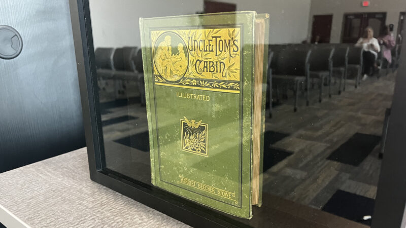 A rare 1880 copy of Uncle Tom's Cabin inside a glass case. It has a green cover with a yellow design around the title and an emblem of a bird diving toward a tree limb intertwined with chains.