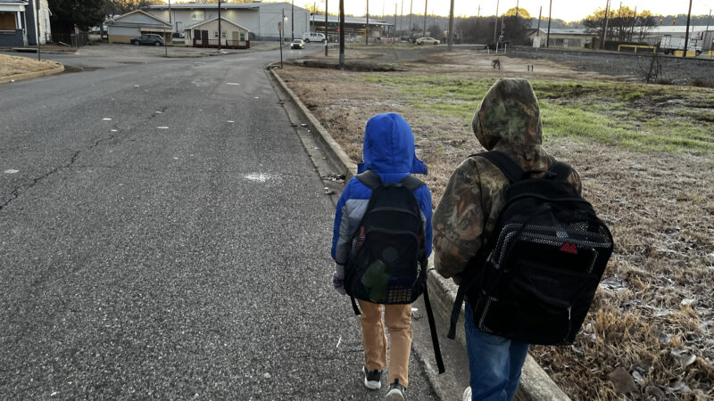 Two young children walk on a road with no sidewalks near a train track. A car drives toward them in the distance.