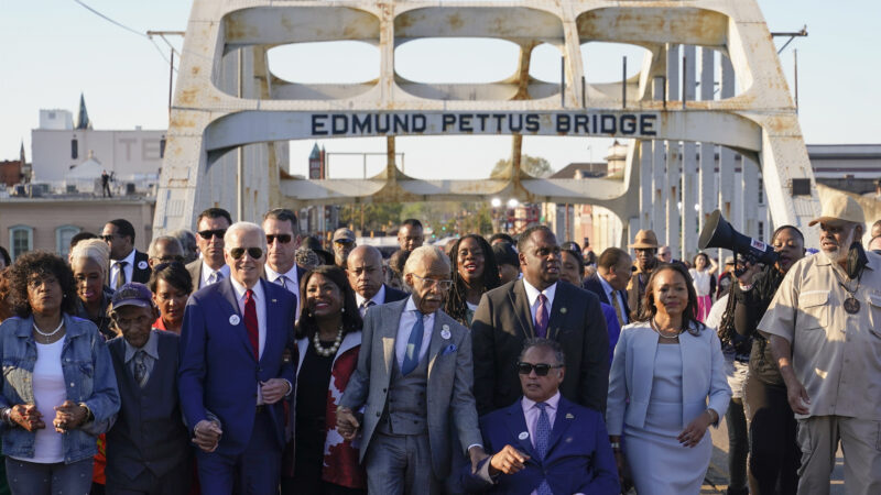 President Joe Biden walks across the Edmund Pettus Bridge in Selma, Alabama next to a large crowd including politicians Terri Sewell and Rev. Al Sharpton.