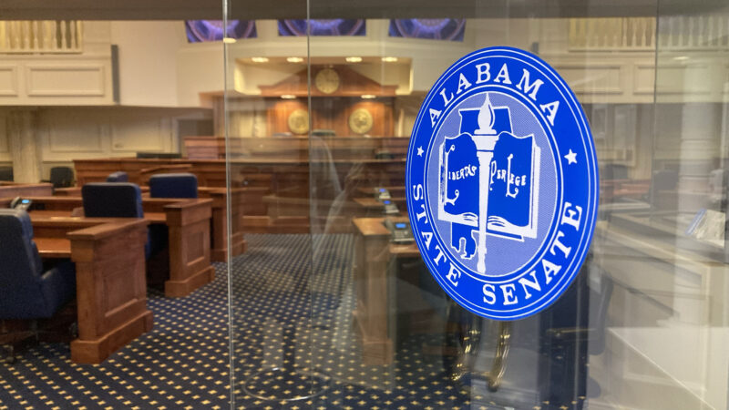 A glass door opens to an empty senate chamber. A blue circular seal on the door shows a design of a book, a torch and the state of Alabama with the text "Alabama State Senate."