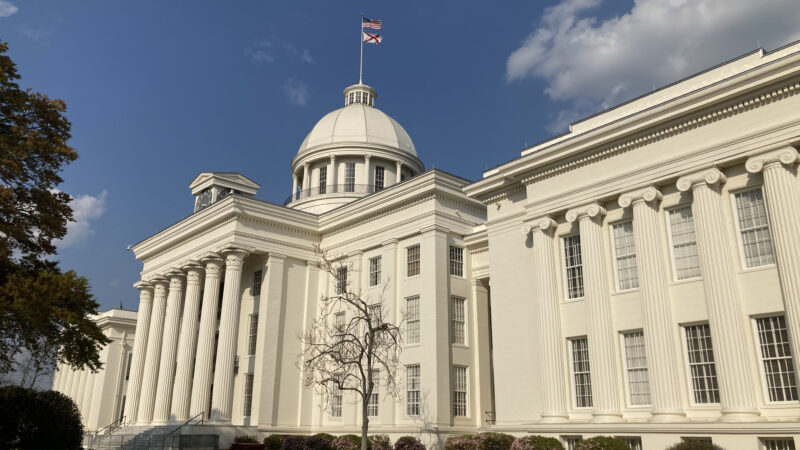 The outside of the Alabama State Capitol building on a sunny day. The Alabama flag and American flag fly at the top of the capitol building's dome.