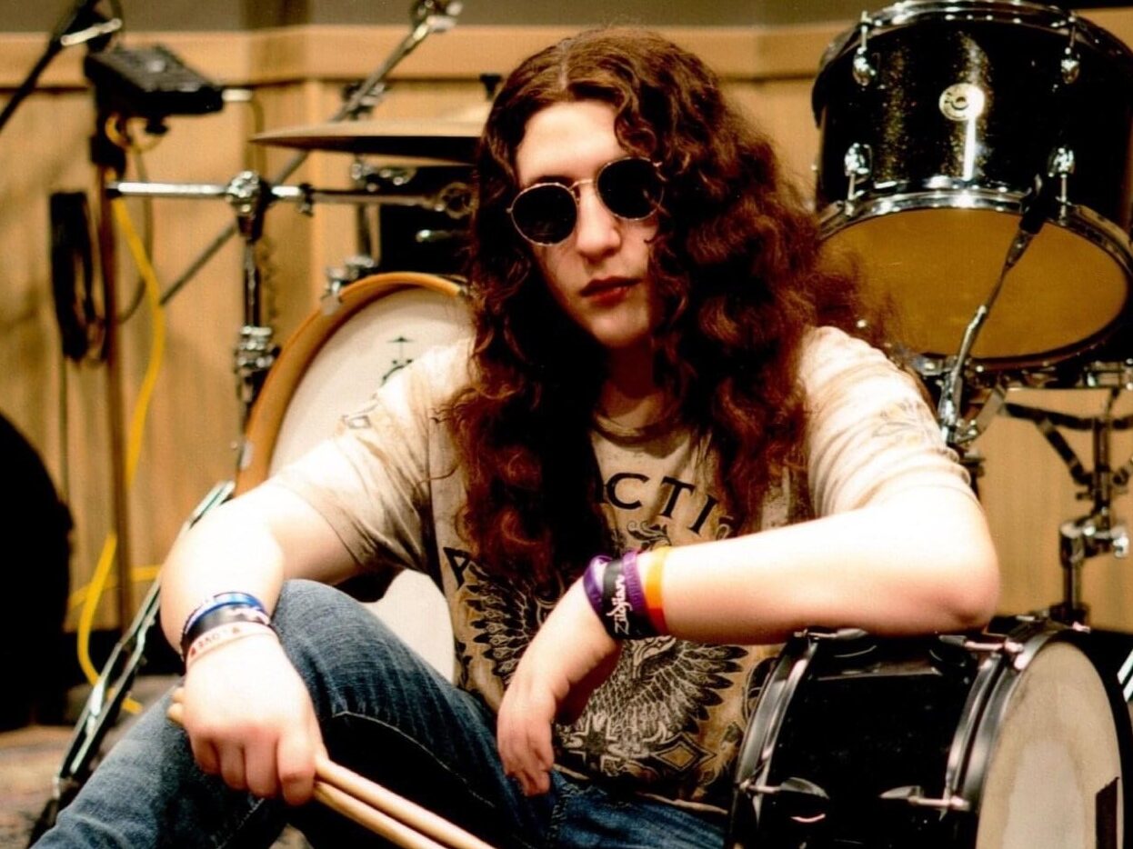 Boy with long hair sits next to drum set