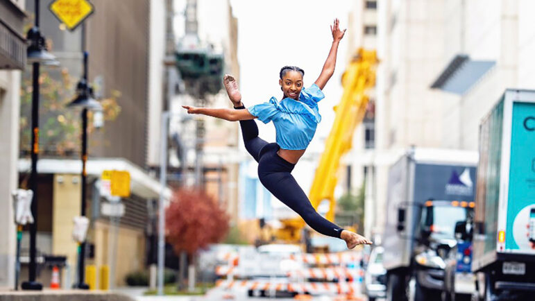 A woman in midair holding a ballet pose in the middle of a busy street.