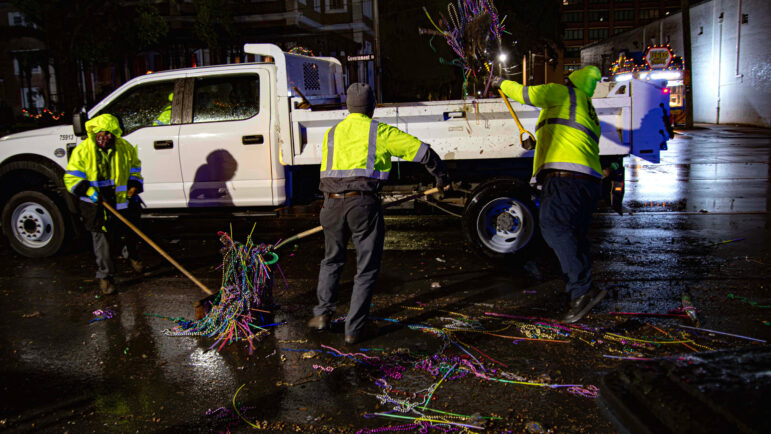Mobile city workers shovel pounds of Mardi Gras beads into the back of a truck.