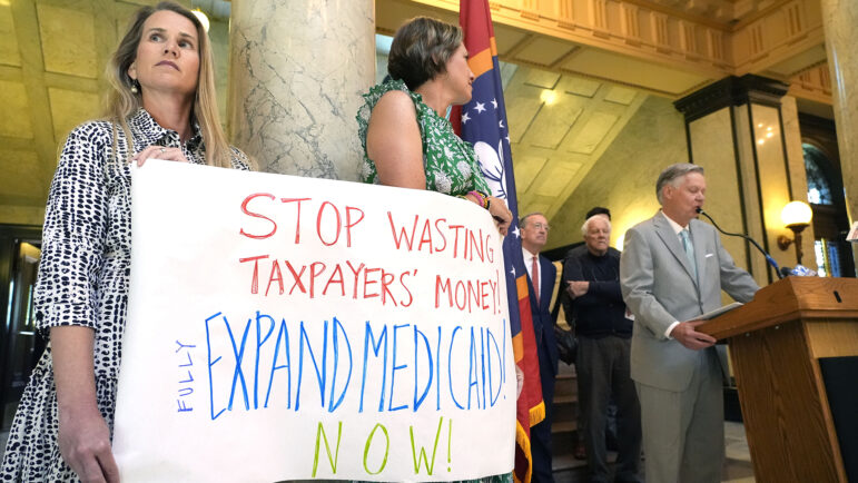 Kathy Knight, left, of Madison, and Kathleen O'Beirne of Ridgeland, hold a sign urging lawmakers to fully fund a Mississippi Medicaid expansion plan, while Jack Reed Jr., a former Tupelo mayor and a long time northeast Mississippi businessman, right, joins a group of small business owners who urged the same during a Tuesday, April 23, 2024, news conference at the state Capitol in Jackson, Miss.