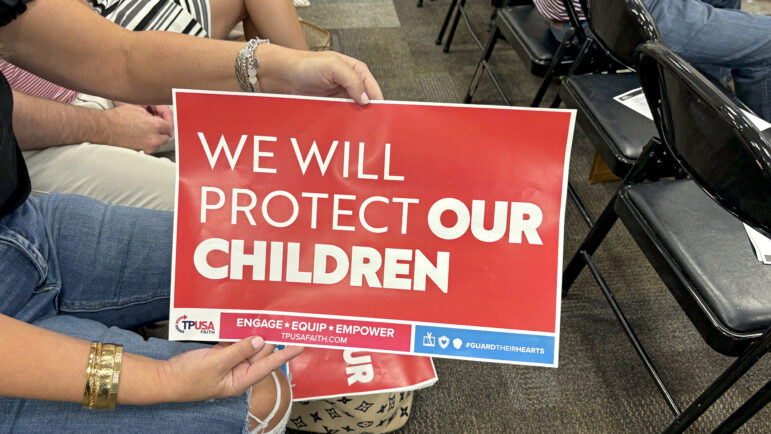 An attendant at the Moms for Liberty town hall meeting about Title IX changes holds a Turning Point USA sign that reads “We will protect our children” on July 1, 2024, in Pelham, Alabama.