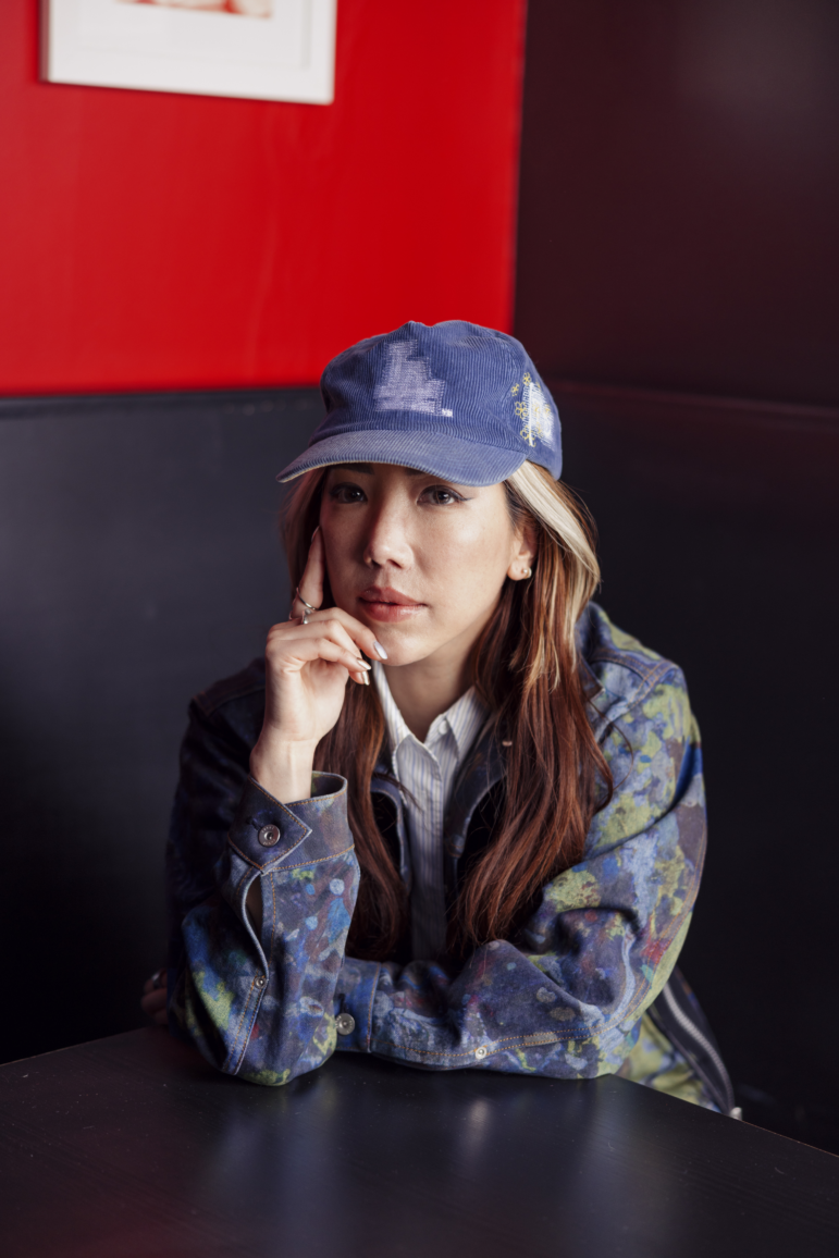 A portrait of Jennifer Lee wearing a blue baseball cap with LA on it. She is wearing a painted denim jacket and is sitting in a booth of a restaurant in the Koreatown neighborhood of Los Angeles.