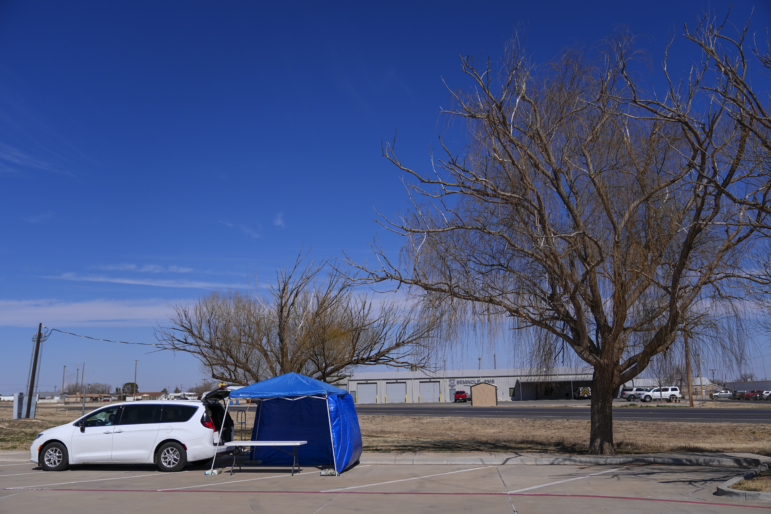 A van is parked next to a blue tent set up as a measles testing location in a parking lot near a tall tree.