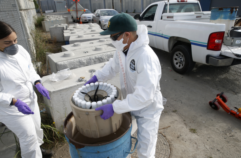 EBMUD wastewater control inspectors Zach Wu and Gabriela Esparza (left) recovers a large bucket of 24 individual bottles containing sewage samples from collection equipment in Oakland, Calif. on Tuesday, July 14, 2020. The samples are sent to a number of labs to analyze for any detection of the COVID-19 coronavirus in the sewage system.