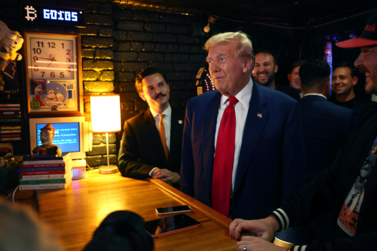 In this photo, then-candidate Donald Trump stands at the wooden bar of Pubkey, a cryptocurrency-themed bar in New York City, on September 18, 2024. He's wearing a blue suit and red tie, and multiple people are standing behind him.