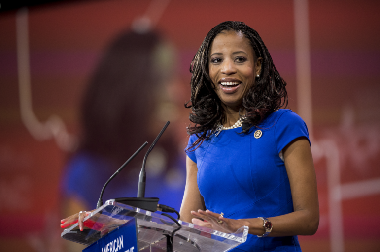 In this 2015 photo, Rep. Mia Love is standing at a lectern with microphones attached to it. She's wearing a blue dress with short sleeves.
