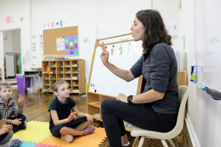 A woman is seated at the front of a classroom with children seated on colorful interlocking-mats on the floor in front of her.
