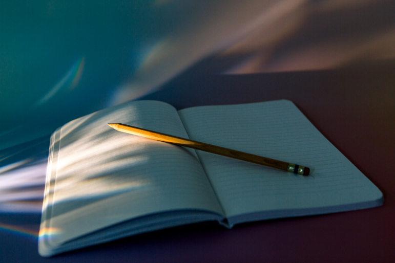 A photograph showing an open, blank notebook with a pencil propped on top of the page laid against a background with a blue-to-pink gradient. Rainbowed light spills onto the page from the left side of the frame.