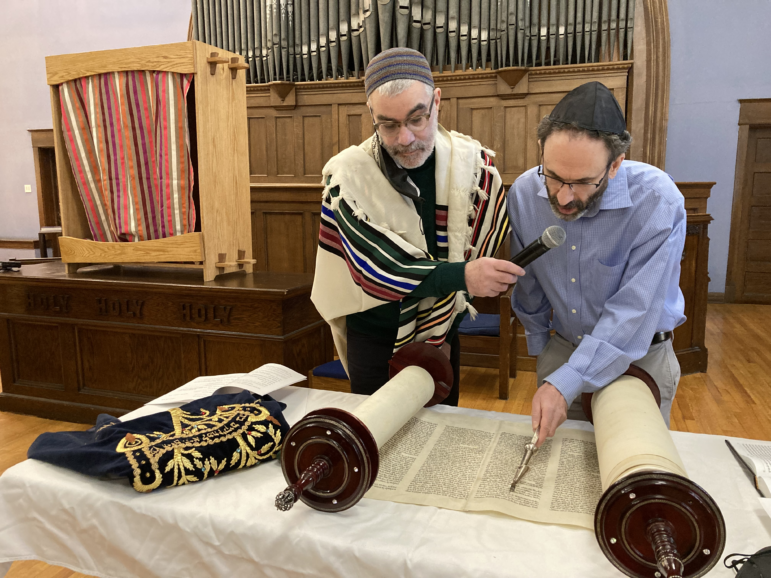 A man reads from the Torah using a traditional pointer to follow the words, while the rabbi looks on, holding a microphone for the reader.