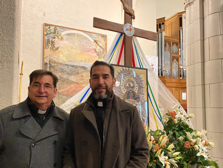 Two Catholic clergy members stand side by side in front of a cross draped with colorful ribbons.