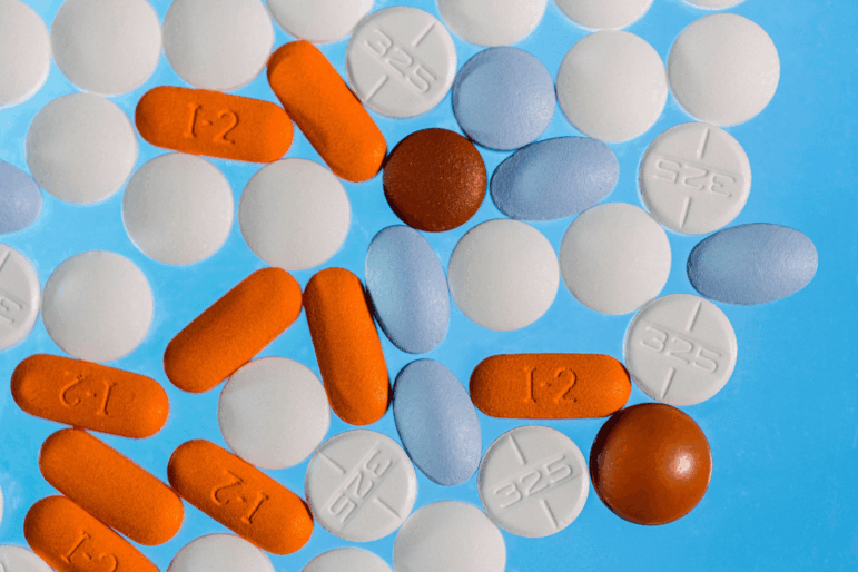 Macro photograph of various over-the-counter pain medicines, including Aleve, Tylenol, Advil and various other NSAIDs and acetaminophen pills, that are a variety of colors, including white, orange, brown and light blue, and that range from circles to elongated ovals, close up on a bright blue backdrop.