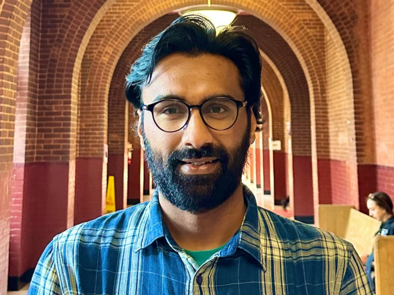 Badar Khan Suri is earing glasses and a blue plaid shirt and standing in a hallway of arches.