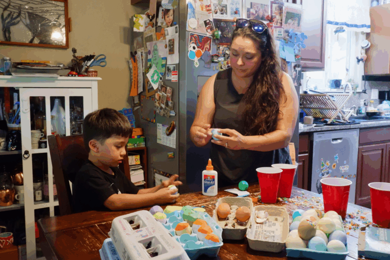 A small boy sits at a table holding an egg, while his mother stands next to him holding one too. The table is covered with colorful eggs, plastic cups and confetti.