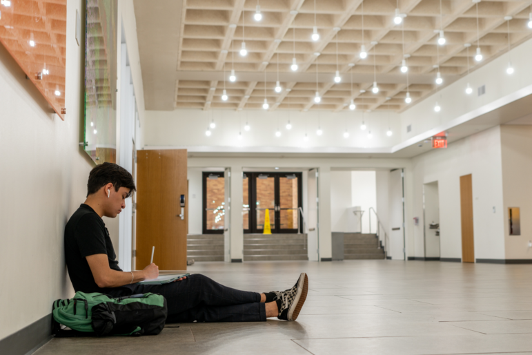 A student sits on the floor in a hallway and writes on a pad on his lap. His backpack is on the floor beside him.