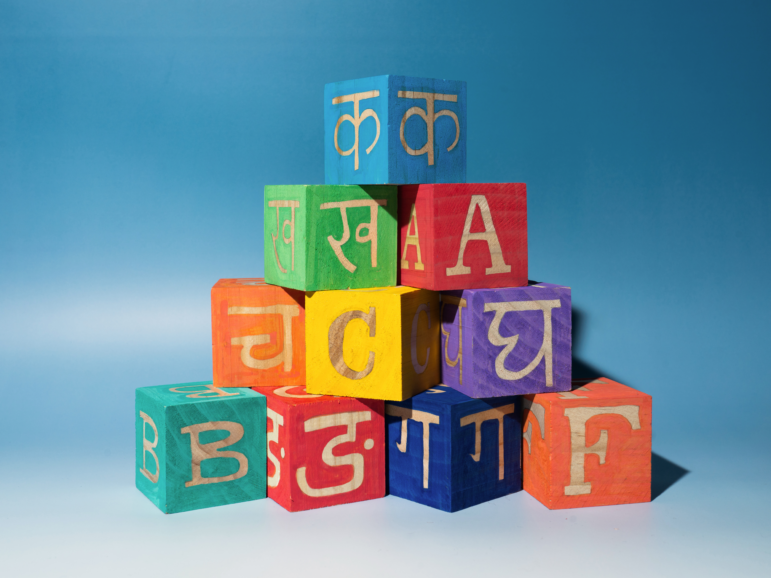 A stack of colorful wooden blocks featuring letters from the Nepali and English alphabet are stacked against a blue backdrop.