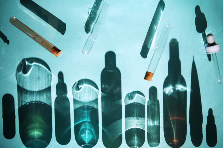 Photograph showing the shadows of several personal care and beauty products on an aqua background. Above the shadows, test tubes and droppers hover, suggesting the ingredients and chemicals that are found in personal care products.
