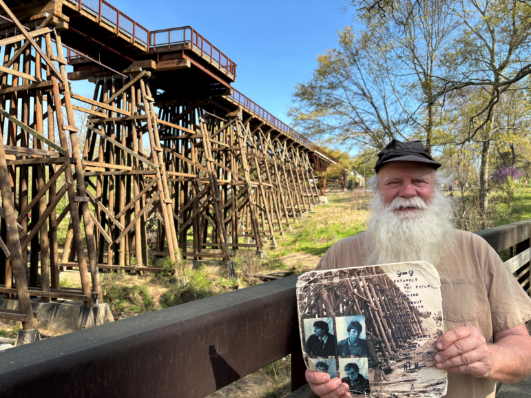 Paul Butchart gives tours of musical landmarks all over Athens, Ga. In his hands is R.E.M.’s 1983 Murmur album, with the original trestle bridge photograph on the back cover. The bridge behind Butchart is a reconstruction, and now part of a walking and biking trail.