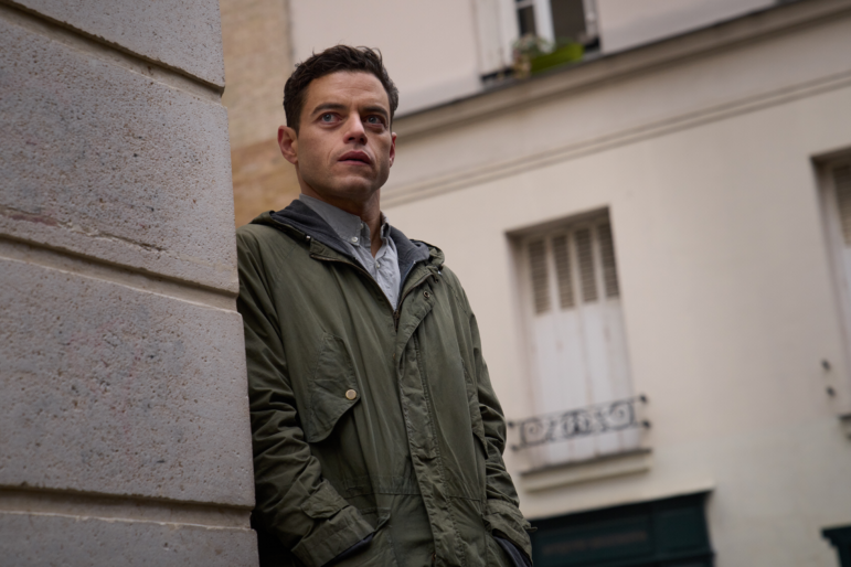 A  man leans against the corner of a building and stares into the distance.