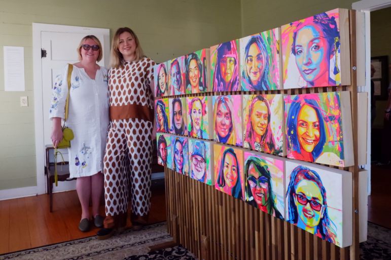 Two women pose next to a piece of art that consists of three rows of brightly colored pop art portraits at the “Many Faces, One Force” exhibit.