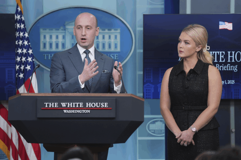 White House Deputy Chief of Staff Stephen Miller gestures behind the podium of the White House briefing room. Press secretary Karoline Leavitt is standing next to him.
