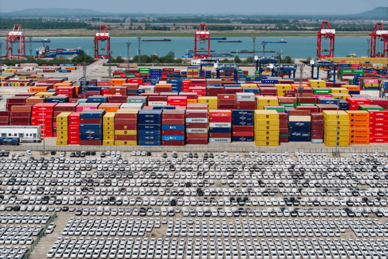 Chinese-made cars are lined up in rows at the port in Nanjing, China. Behind the cars are stacks of cargo shipping containers in a variety of colors — blue, red, yellow, brown, orange.