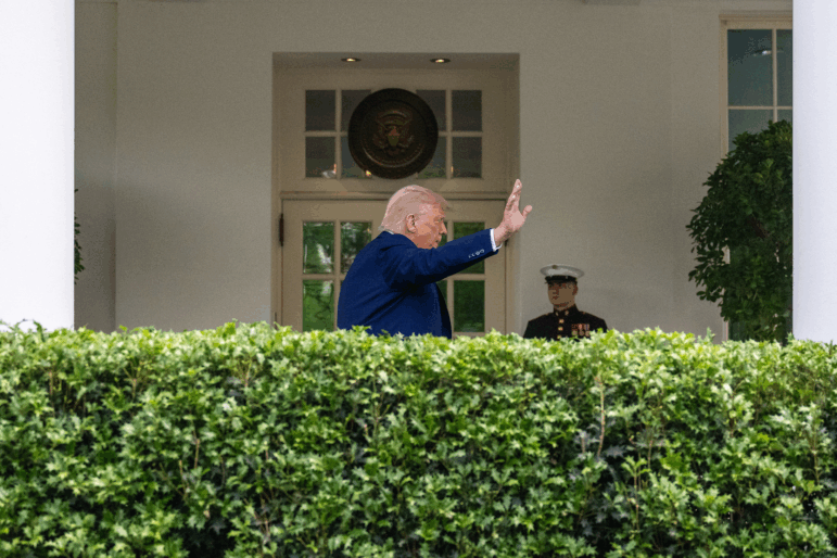 In this photo, President Trump is waving with one hand while outside the White House. A row of hedges is in the foreground.